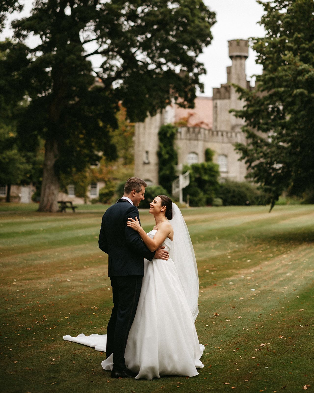 Bride and groom look at each other with so much romance in a lush green yard with castle in the background