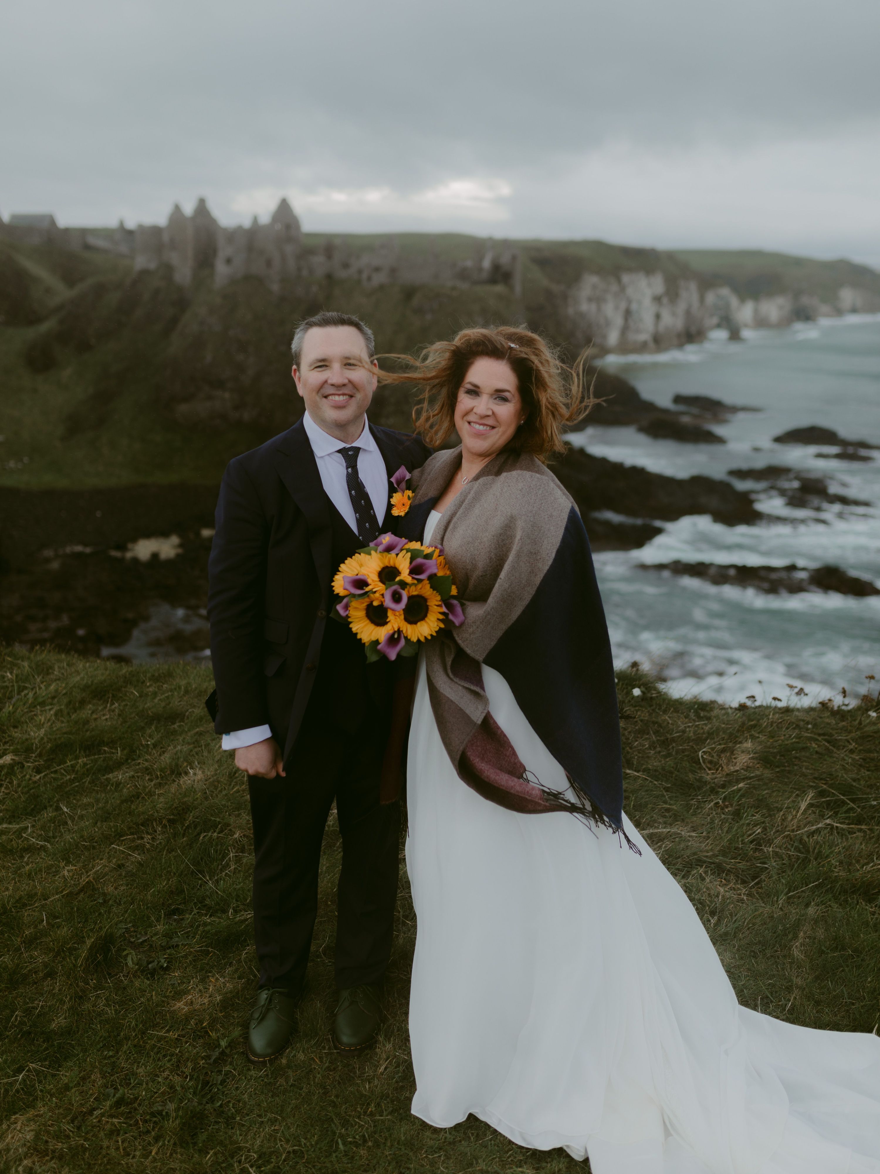 Bride and groom looking at the camera, with bride holding a sunflower bouquet, both atop a cliff after their Irish elopement