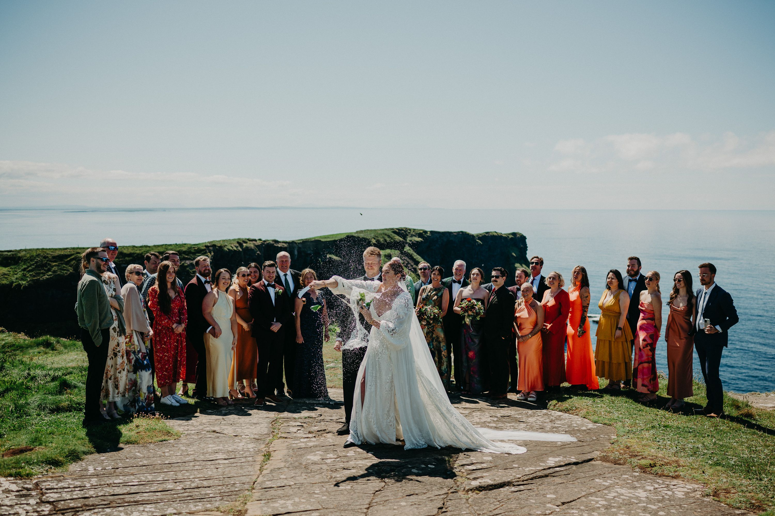 A bride and groom opening a bottle of champagne using a knife while the entire party looks on during an intimate wedding in Ireland