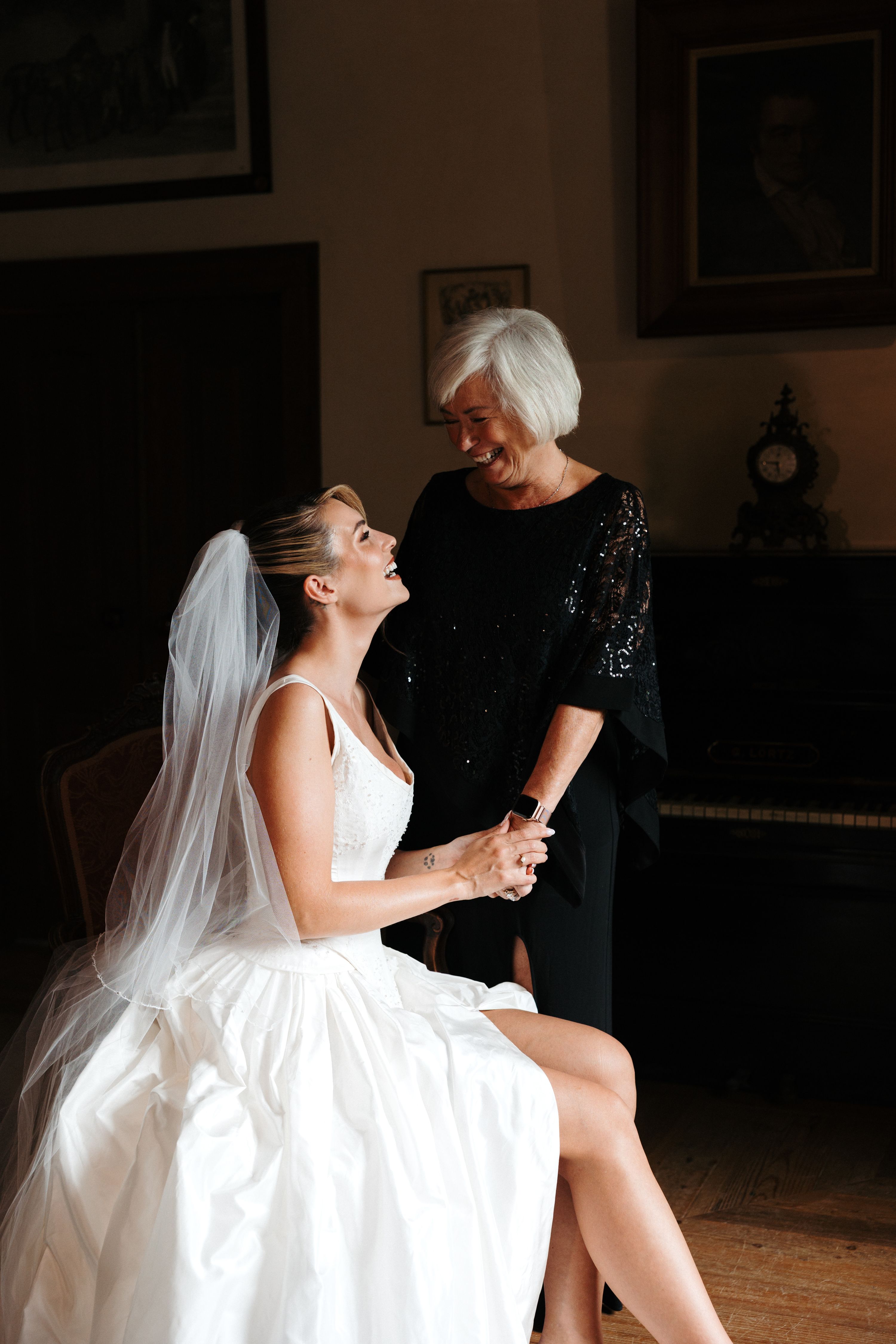 Bride sitting on a chair with her mother happily holding her hand and standing next to her