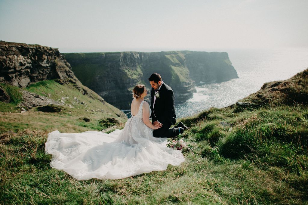 Bride and groom sit atop the meadows overlooking the Cliffs of Moher and the ocean in the background