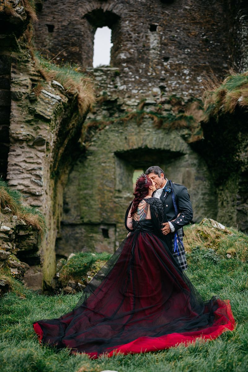 Gothic bride in a black dress and groom in a kilt kissing among castle ruins after eloping in Ireland