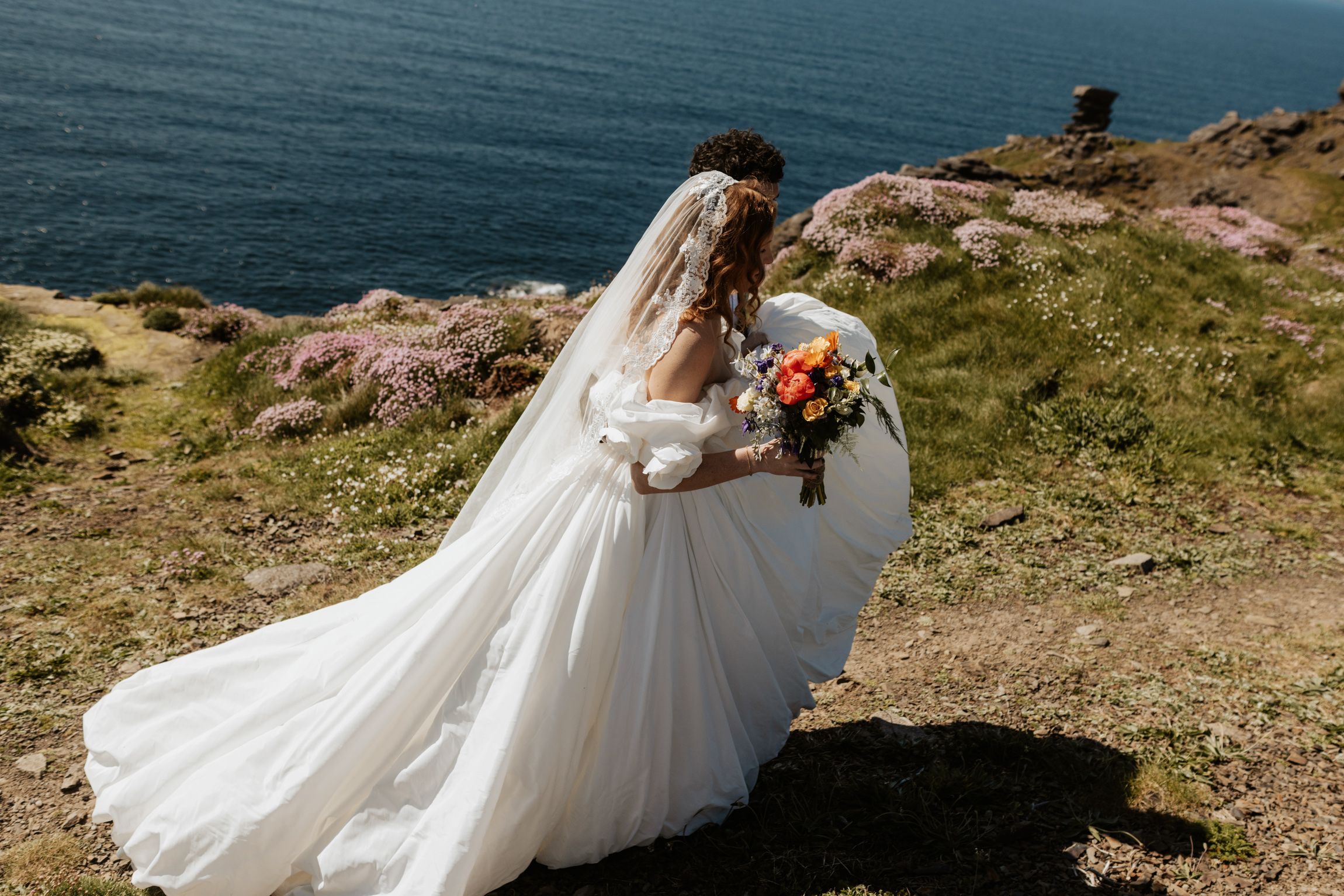 A bride's long lace veil and full gown trail through pink sea thrift on an Irish clifftop above the Atlantic.