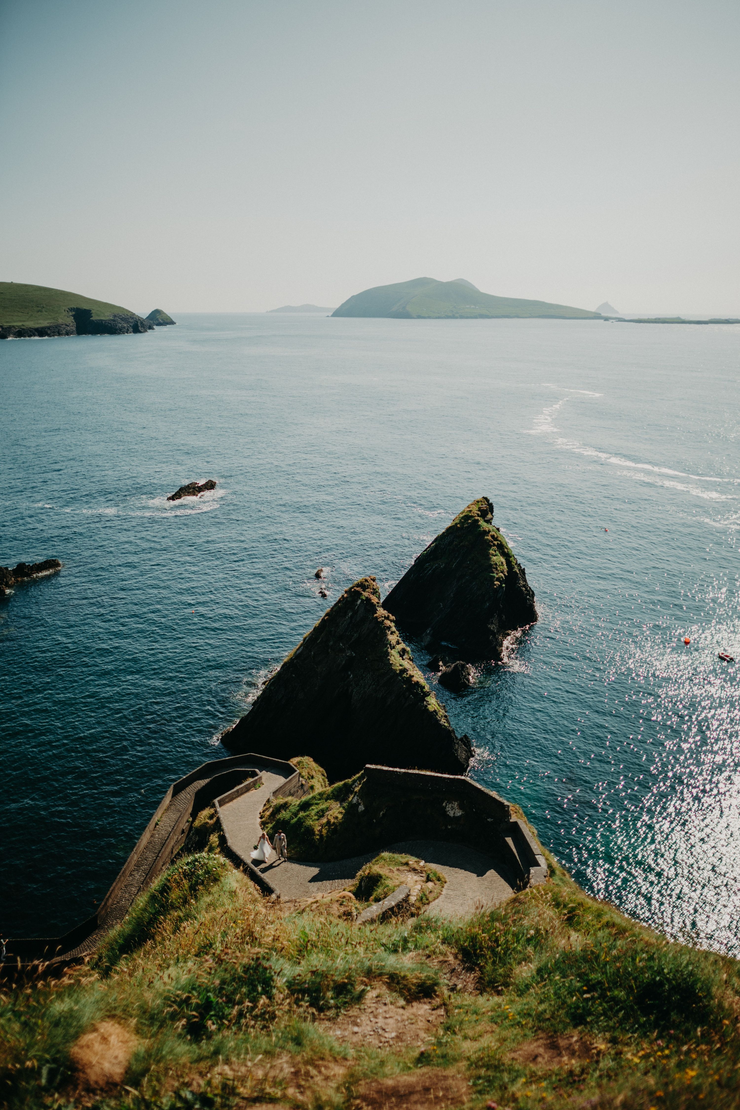 Aerial view of Dunquin Pier's zigzag path winding down to the sea, a couple tiny on the ramp and the Blasket Islands on the horizon.