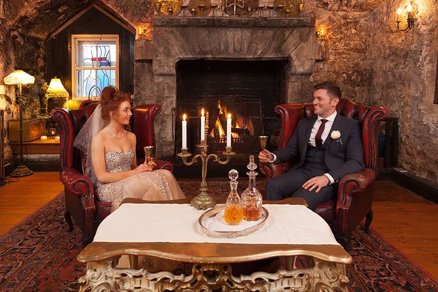 Bride and groom raise a toast in red leather wingback chairs beside the lit medieval fireplace of the Tower Room.