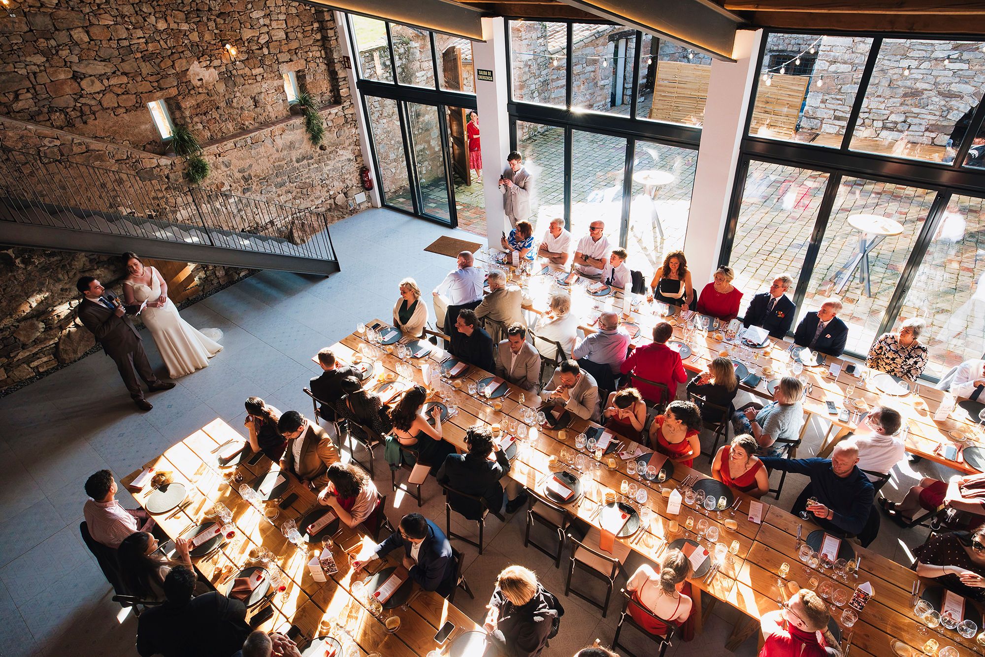 An indoor wedding reception in Barcelona with the newlyweds in front and their guests seated on chairs next to long tables