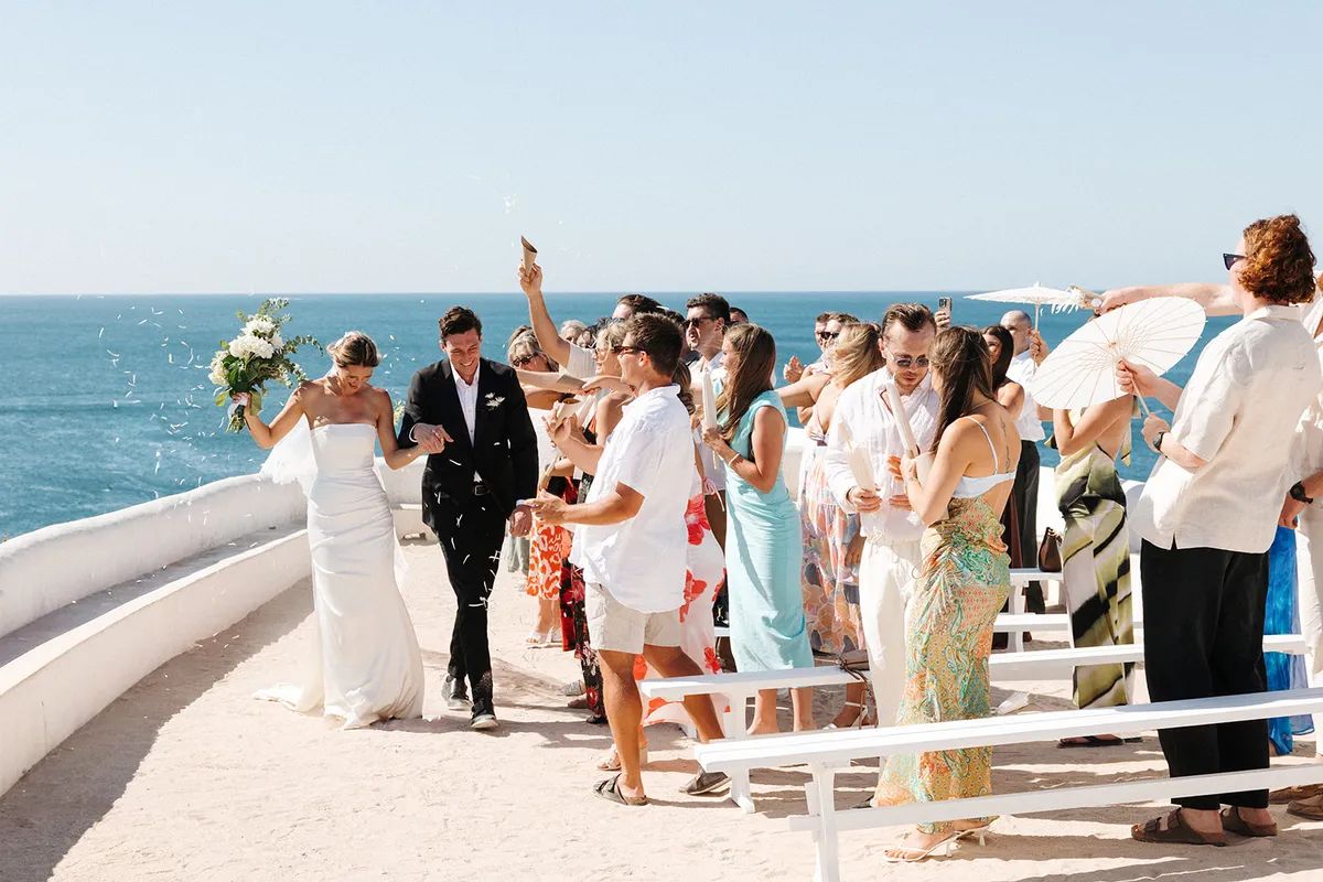 Newlyweds walk through a shower of confetti along a whitewashed terrace above Portugal's Atlantic coast, guests cheering with a parasol.