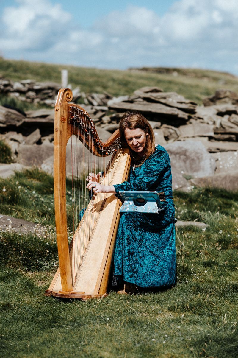 A harpist in a grassy field wearing a blue green velvet dress playing a wooden harp for a destination wedding in Ireland