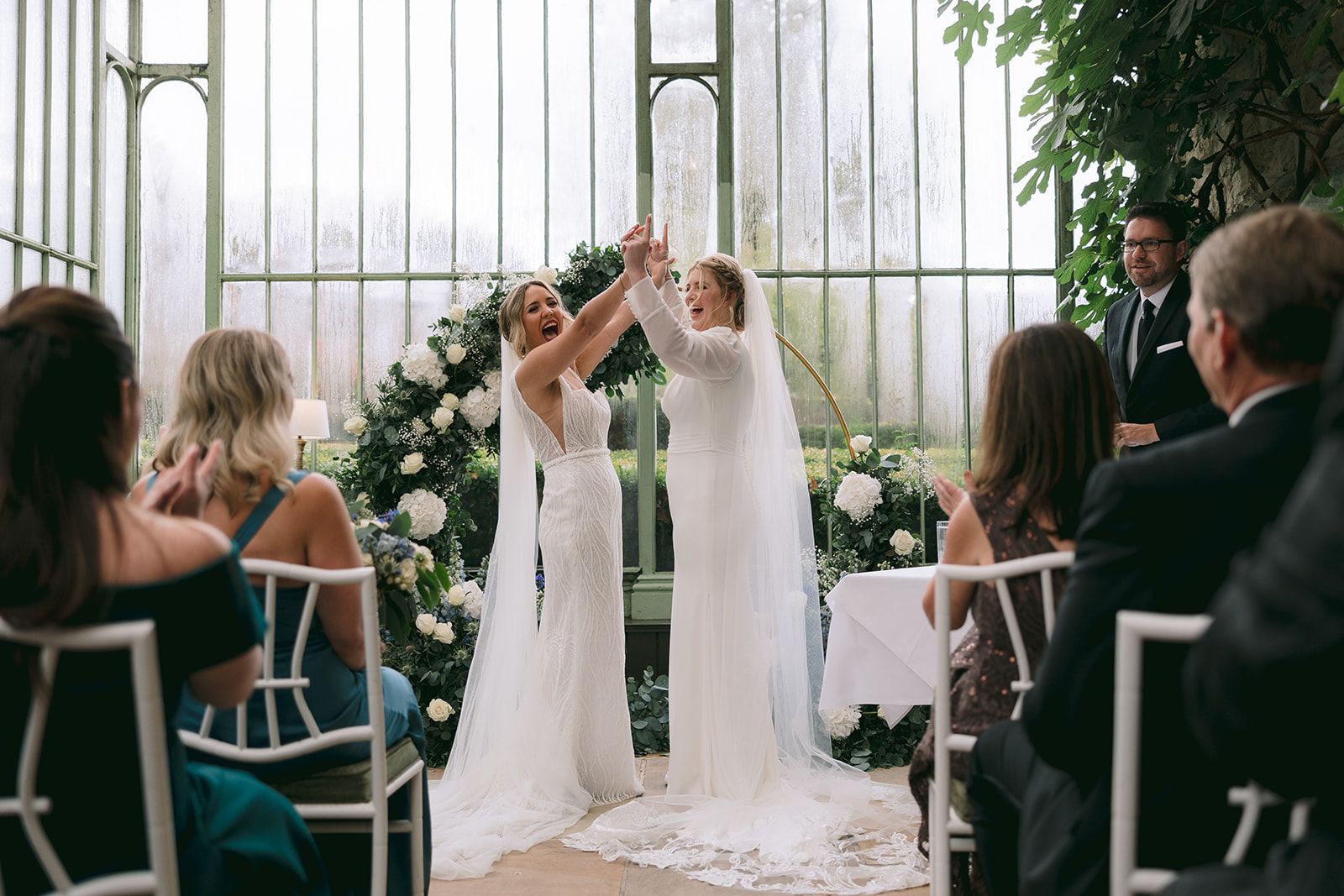 Two brides happily holding hands in front of their guests in a glass-walled ceremony space during their micro wedding in Ireland