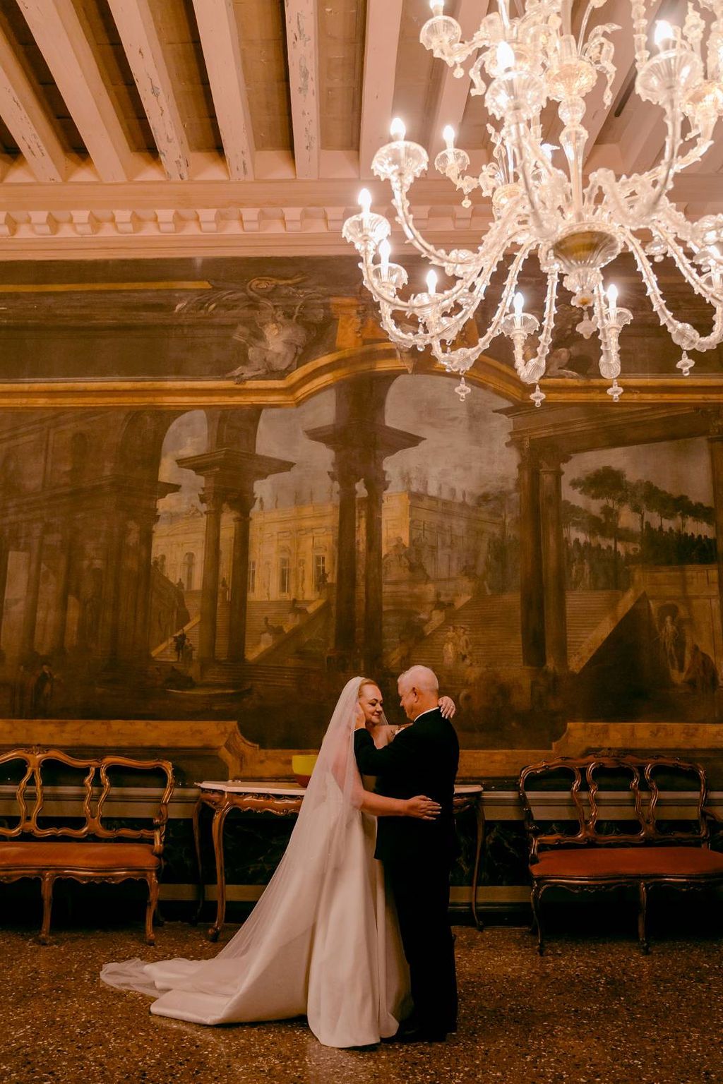 Bride and groom hugging with old paintings in the background and an exquisite chandelier during their vow renewal in Italy