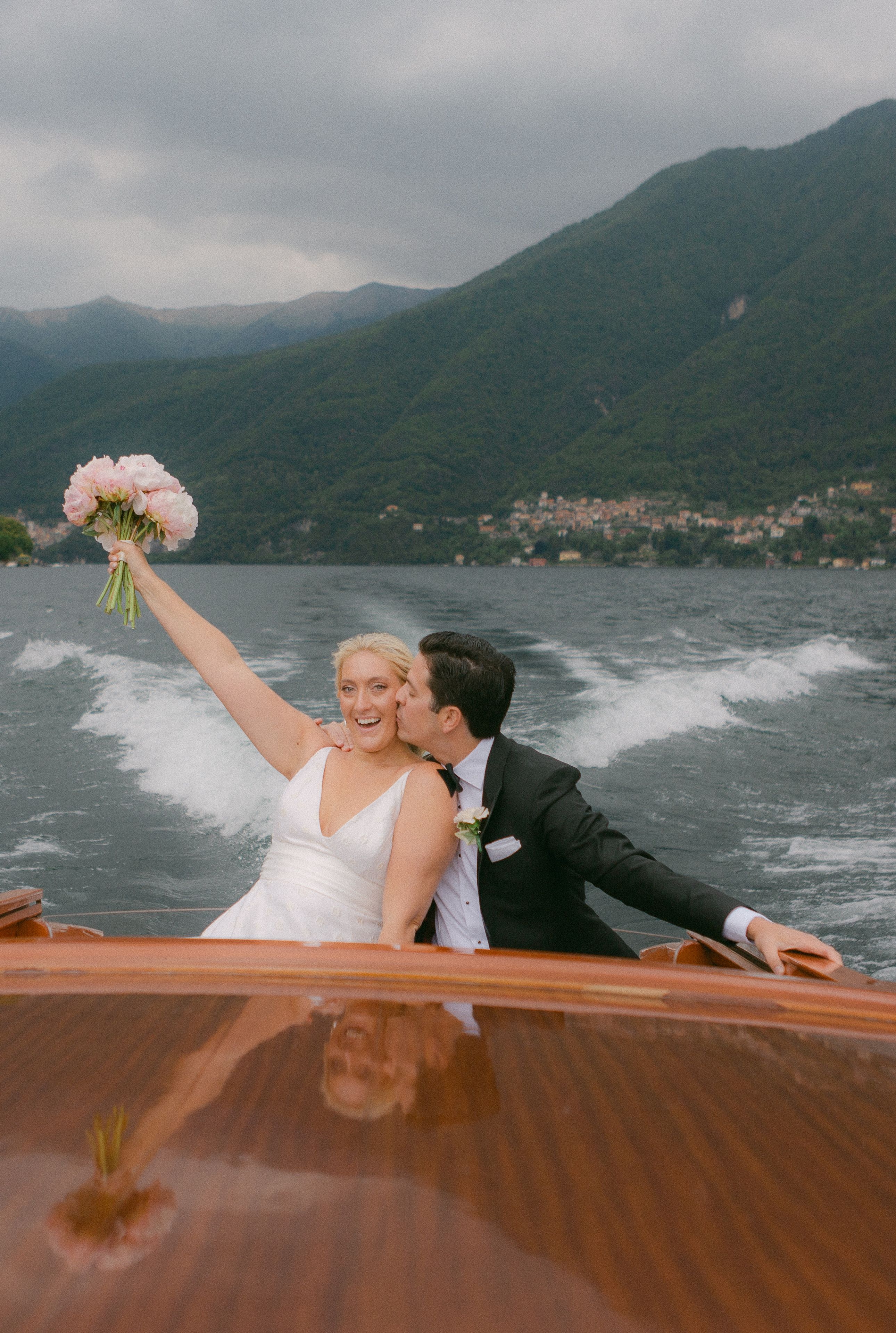 Newlyweds on a boat sailing atop Lake Como with views of the Como mountains and the lake at the back