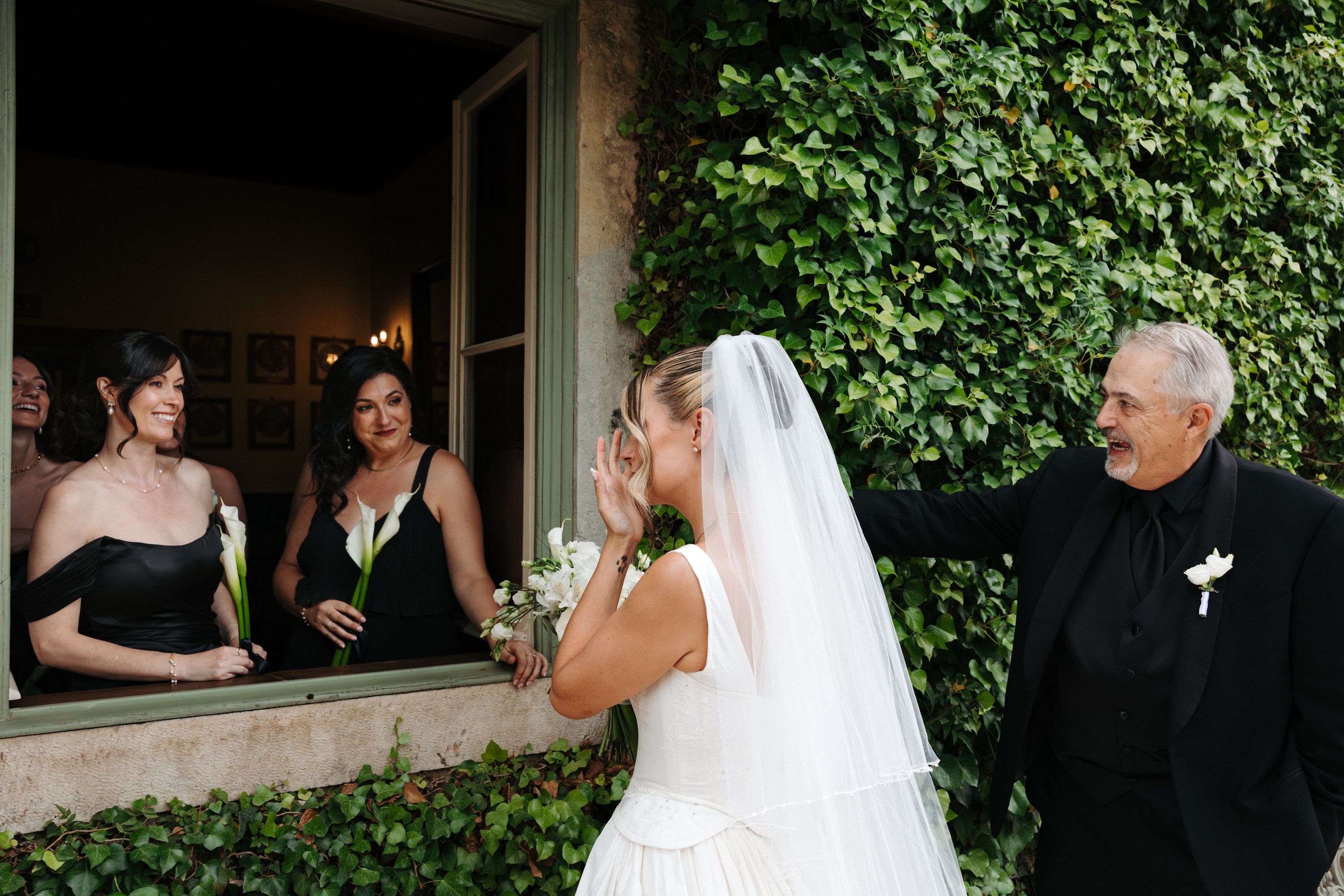 A bride wiping away tears as her father and her family watches her from behind a window