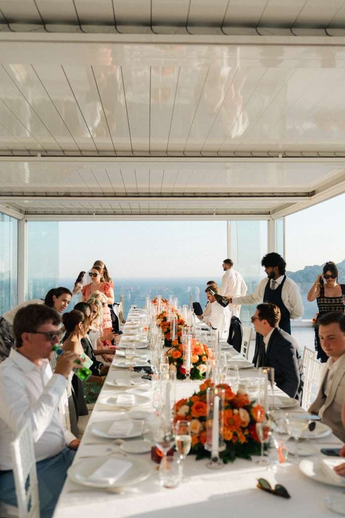Guests are seated on a long table, and are drinking wine during a reception of an Italian intimate wedding