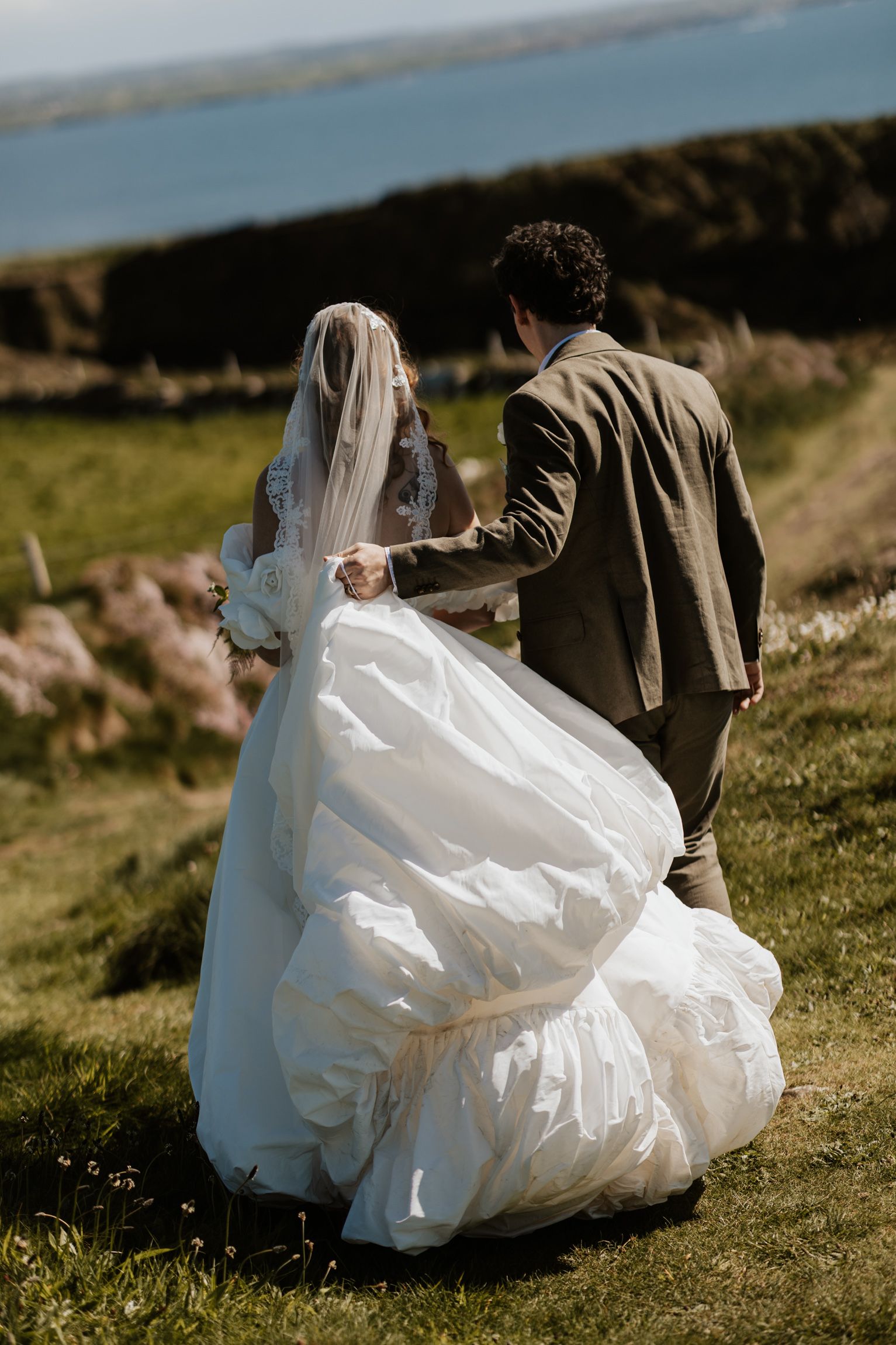 Her groom leads the way as the bride lifts her voluminous train across clifftop grass above the Atlantic near Hag's Head, Ireland.