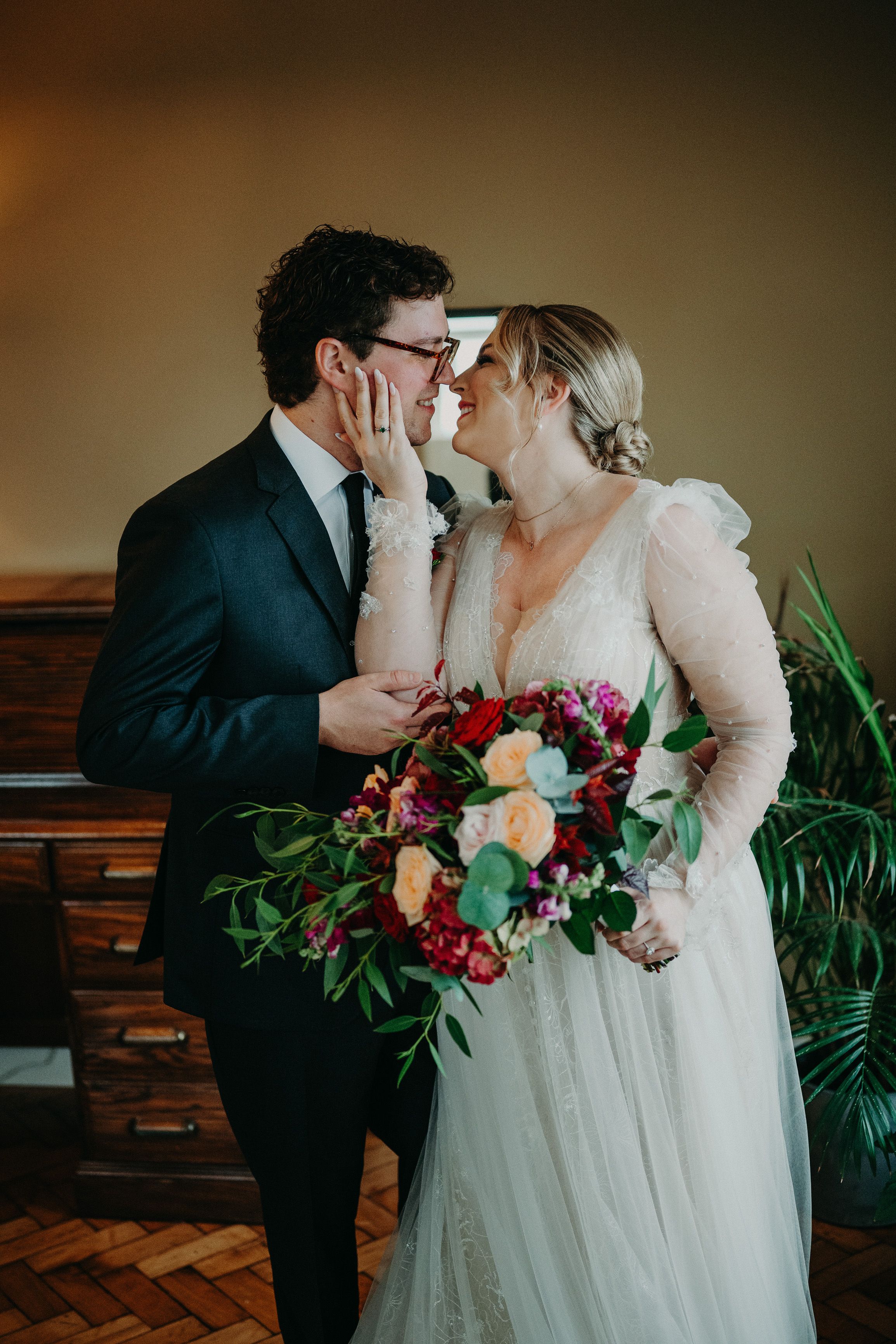 Bride and groom nearly kissing indoors, bride in a sheer long-sleeved gown holding a vibrant bouquet of red and peach roses