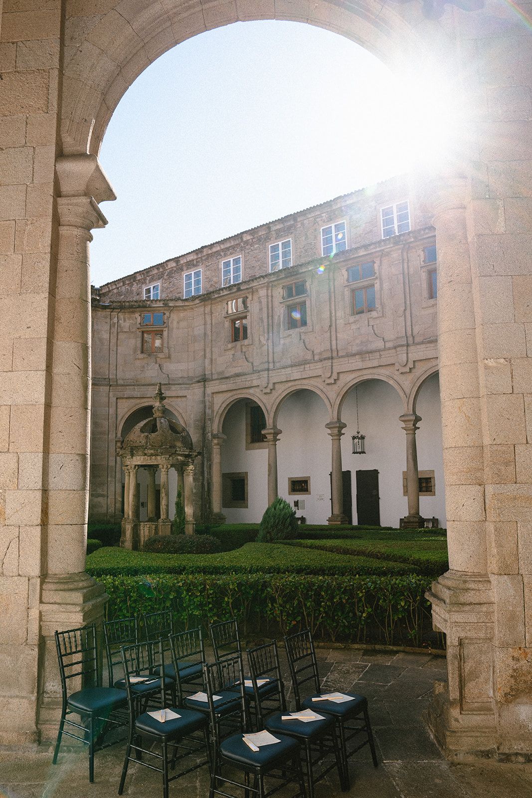 Black chiavari chairs set for a cloister wedding ceremony, sun flaring over the parterre and well-pavilion of the Parador de Santiago.