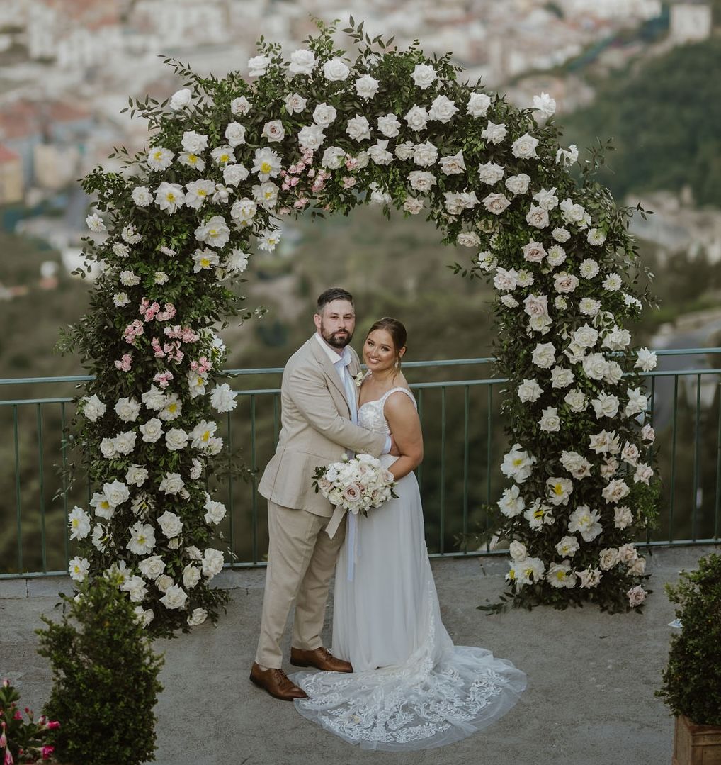 Newlyweds smiling at the camera during their destination elopement in Italy with a floral arch and Amalfi in the background