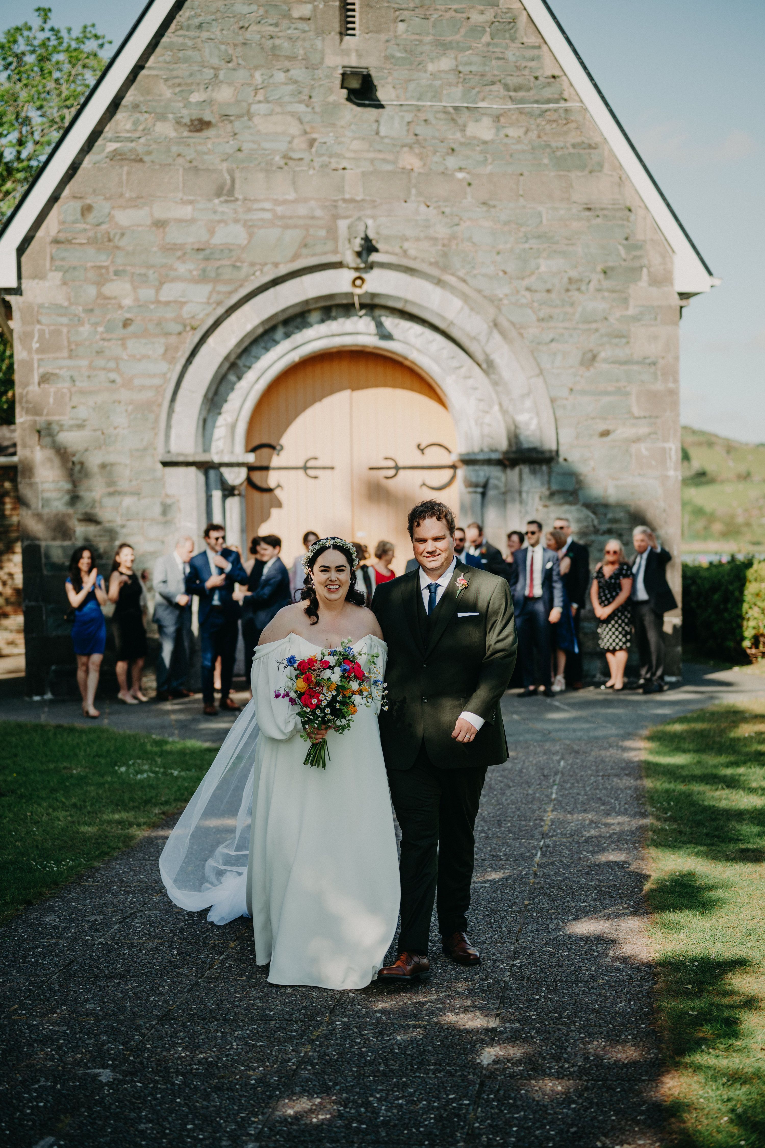 Newlyweds in front of a chapel with a closed wooden door and their guests behind them during their Irish destination wedding