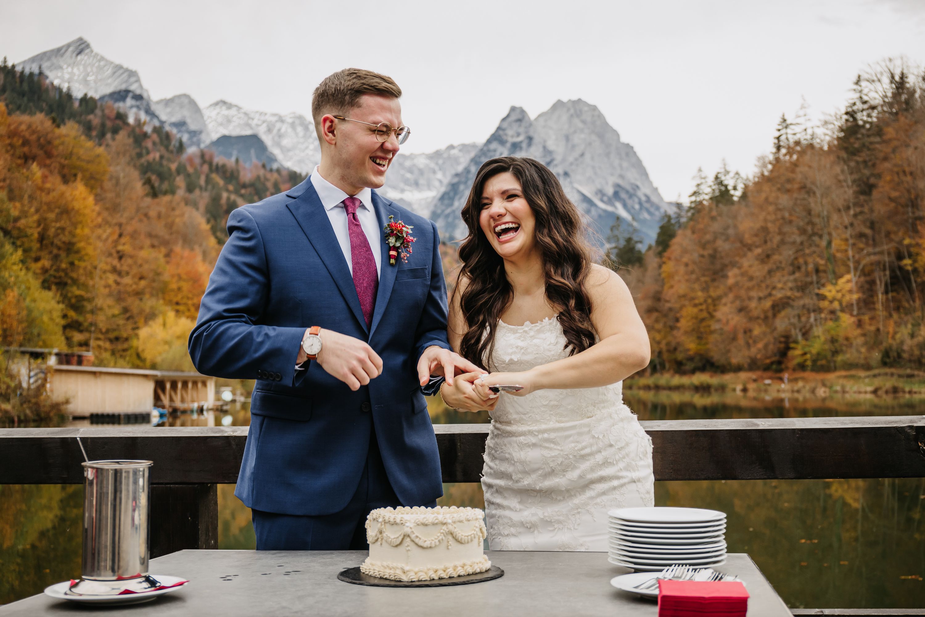 Newlyweds laughing and about to slice a cake with autumn alpine backdrop.