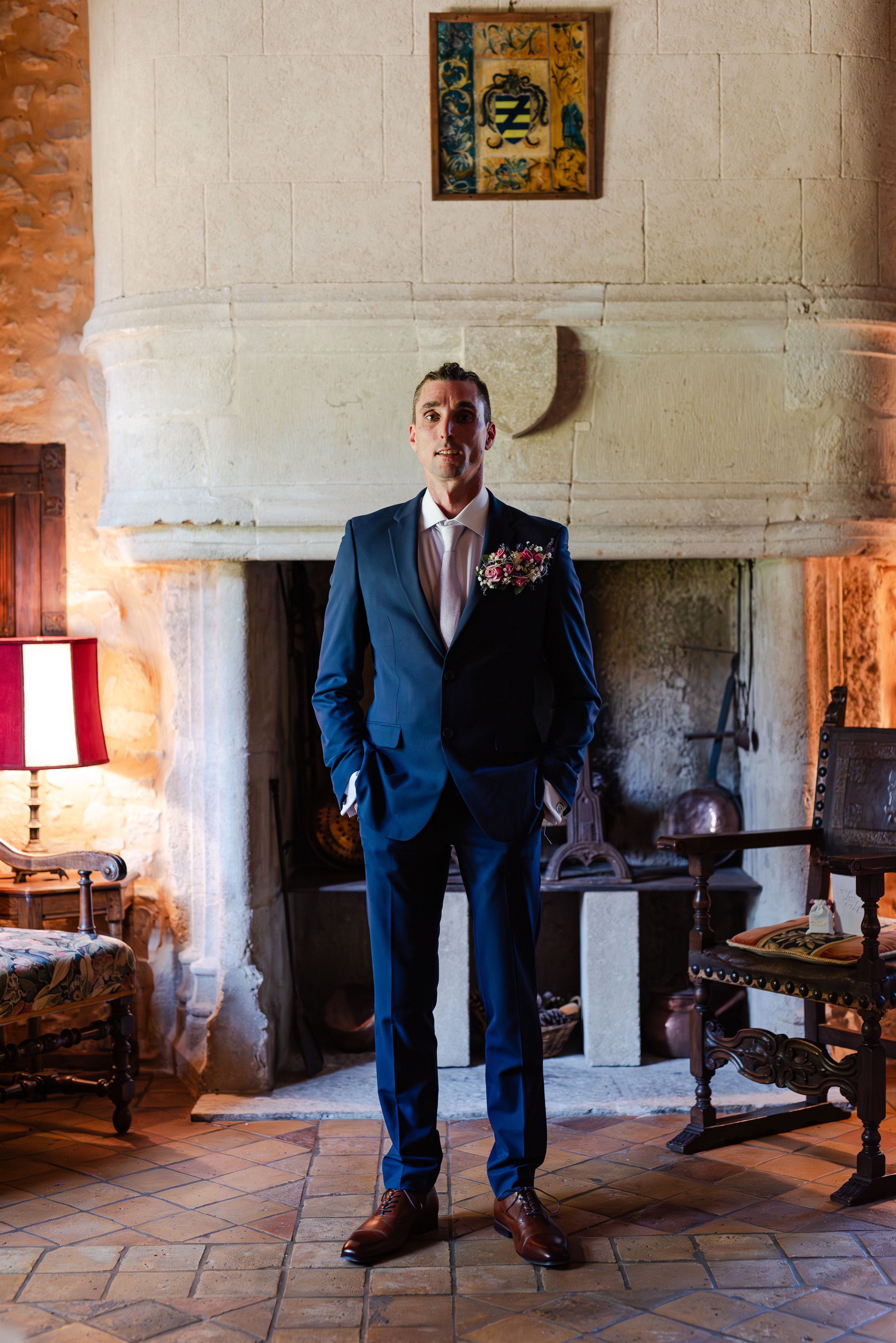 A groom wearing a blue suit with a boutonniere and tie inside a chateau before his small wedding in France