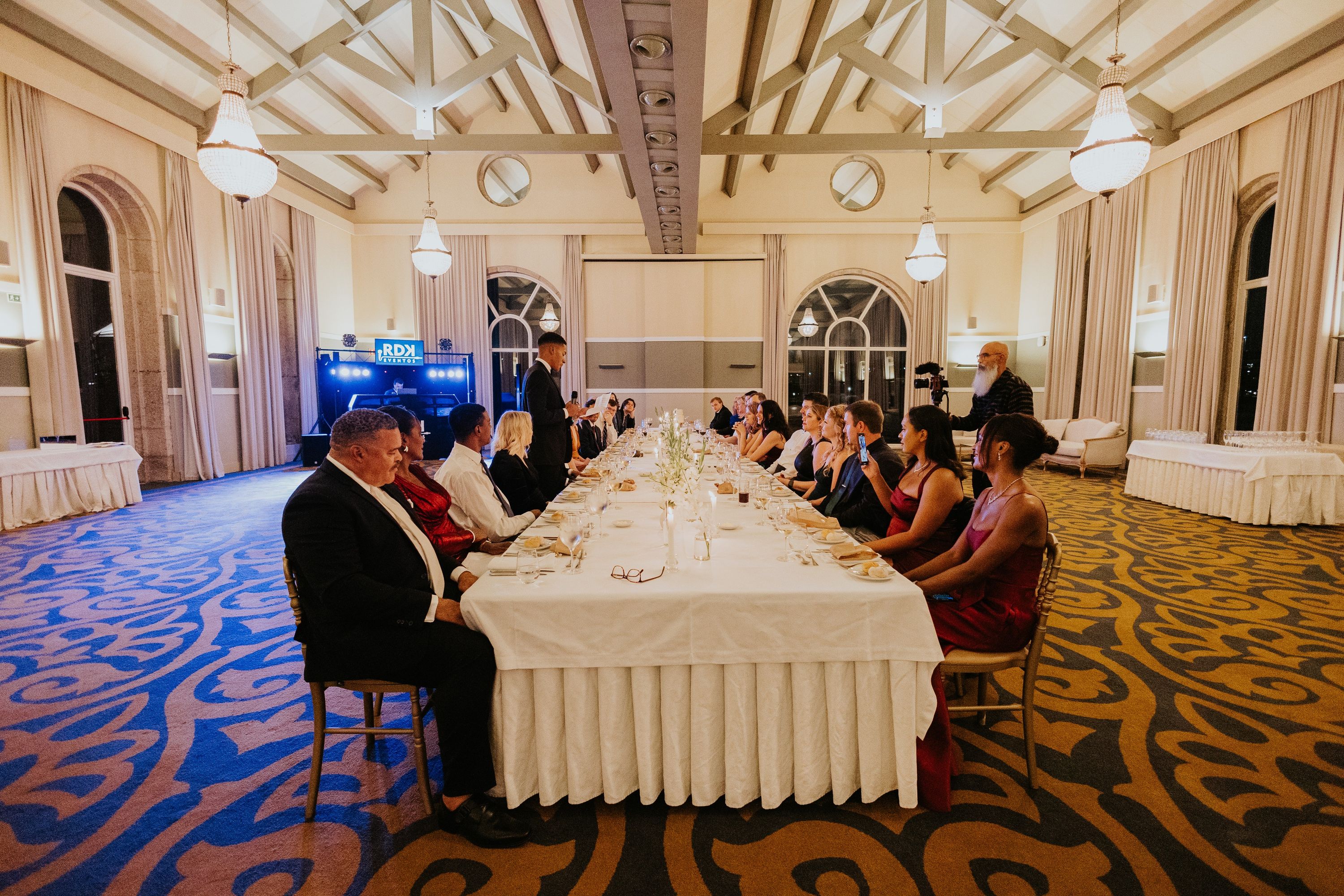 A long white table with guests seated on both sides, with a ceiling adorned with wooden beams and lights