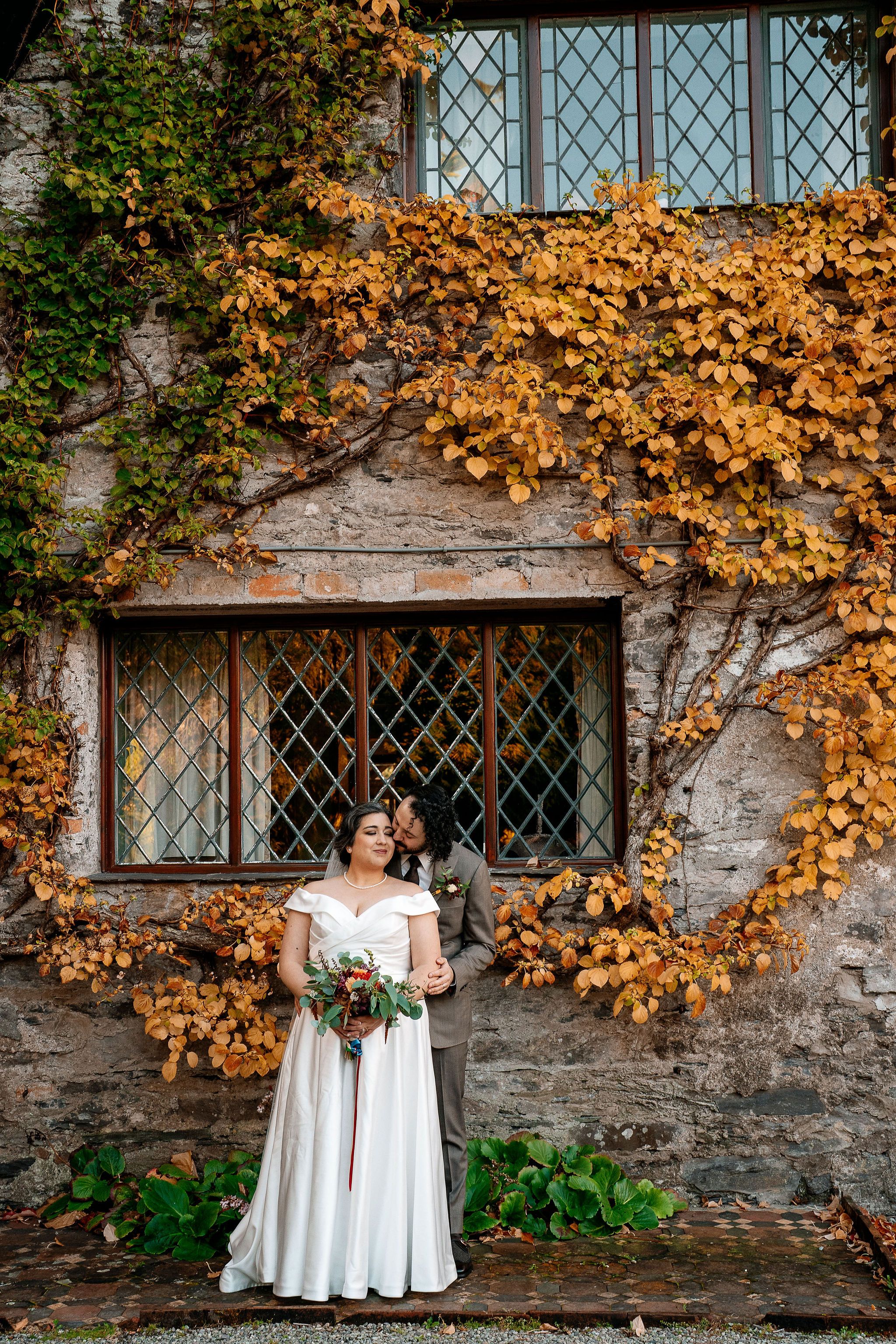 Bride and groom posing in front of a cottage with crawling ivy in autumn blooms during their elopement in Ireland