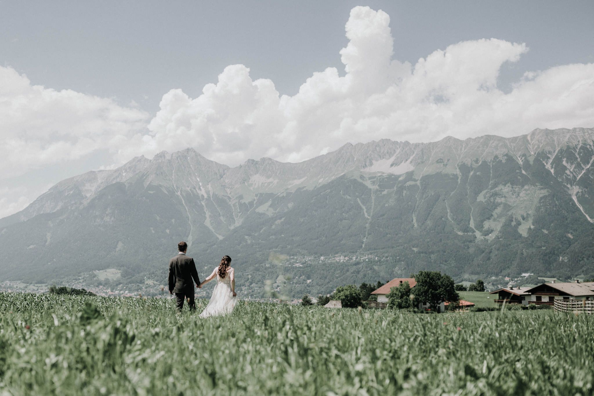Couple walking hand in hand through farmland beneath a long Alpine ridge