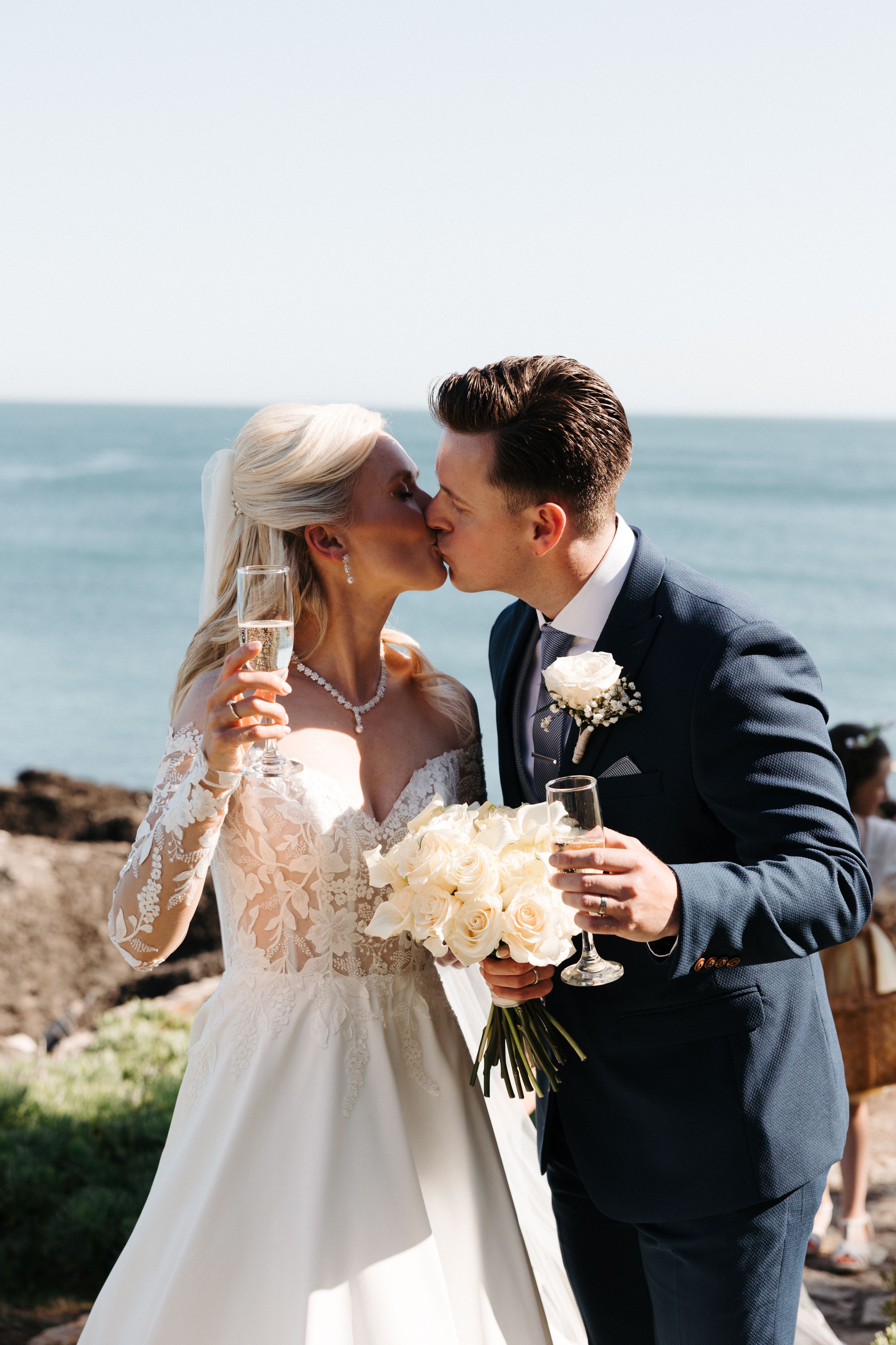 Bride and groom kissing while holding champagne glasses with a beach background during their destination wedding in Portugal