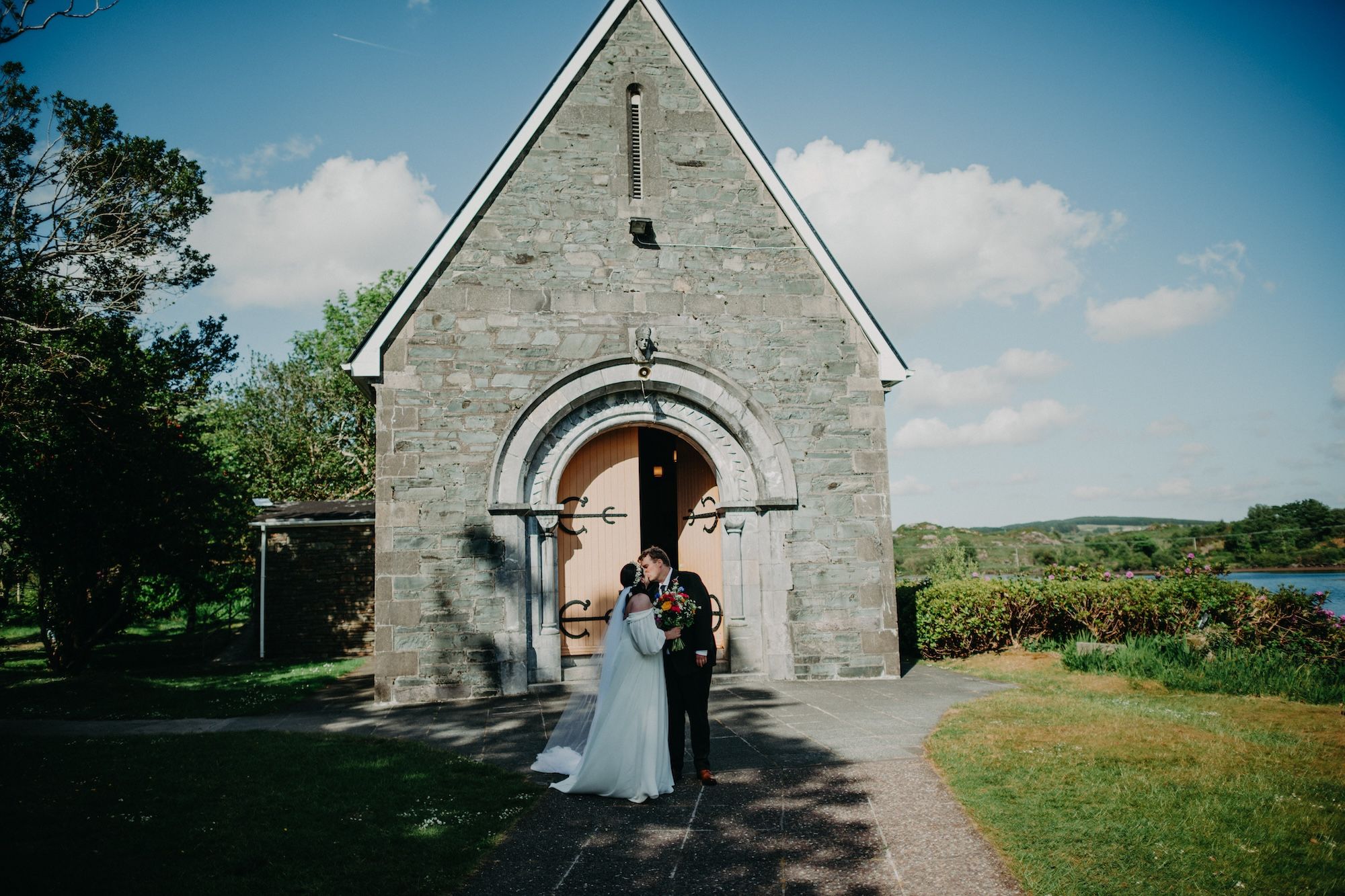 Bride and groom stand in front of a quiant chapel set amidst lush Irish nature during their small wedding in Ireland