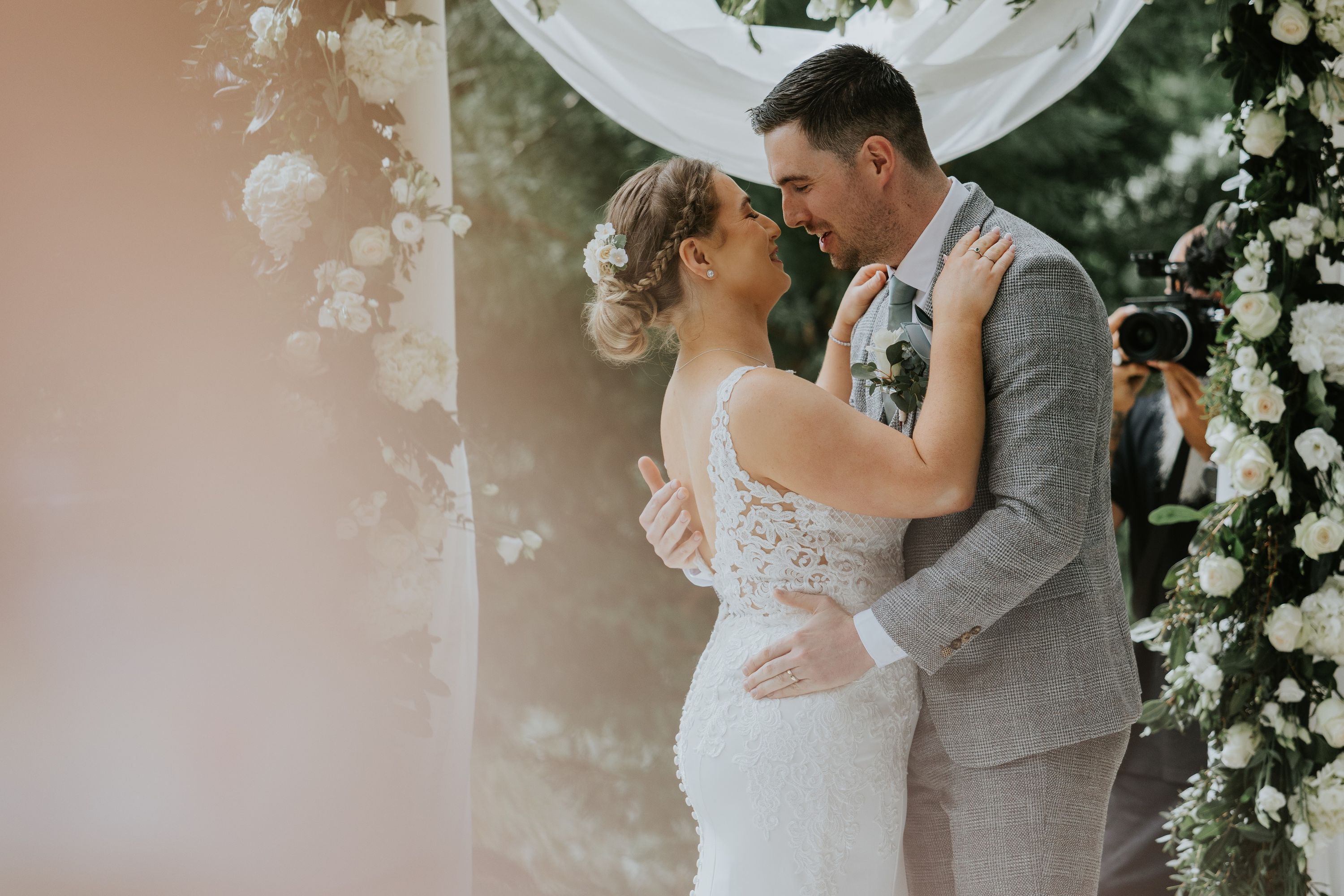 Bride and groom romantically hugging and looking at each other with a floral arch and a white lacey cloth in the background